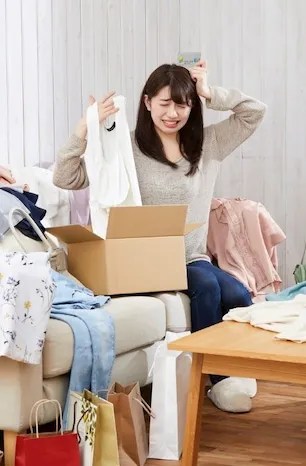一名女性坐在沙發上，手中拿著衣物，看起來感到困惑和焦慮，周圍有許多未整理的包裹和衣物，似乎在處理過多的購物物品。